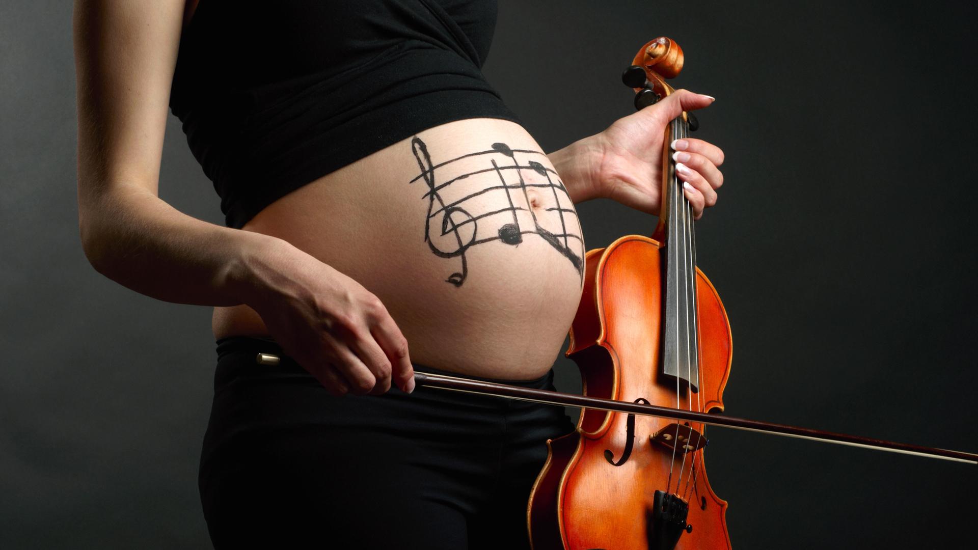 Blick auf einen Schwangerschaftsbauch, auf dem Notenlinien aufgemalt sind, wobei die junge Frau eine Violine und einen Bogen in der Hand hält.