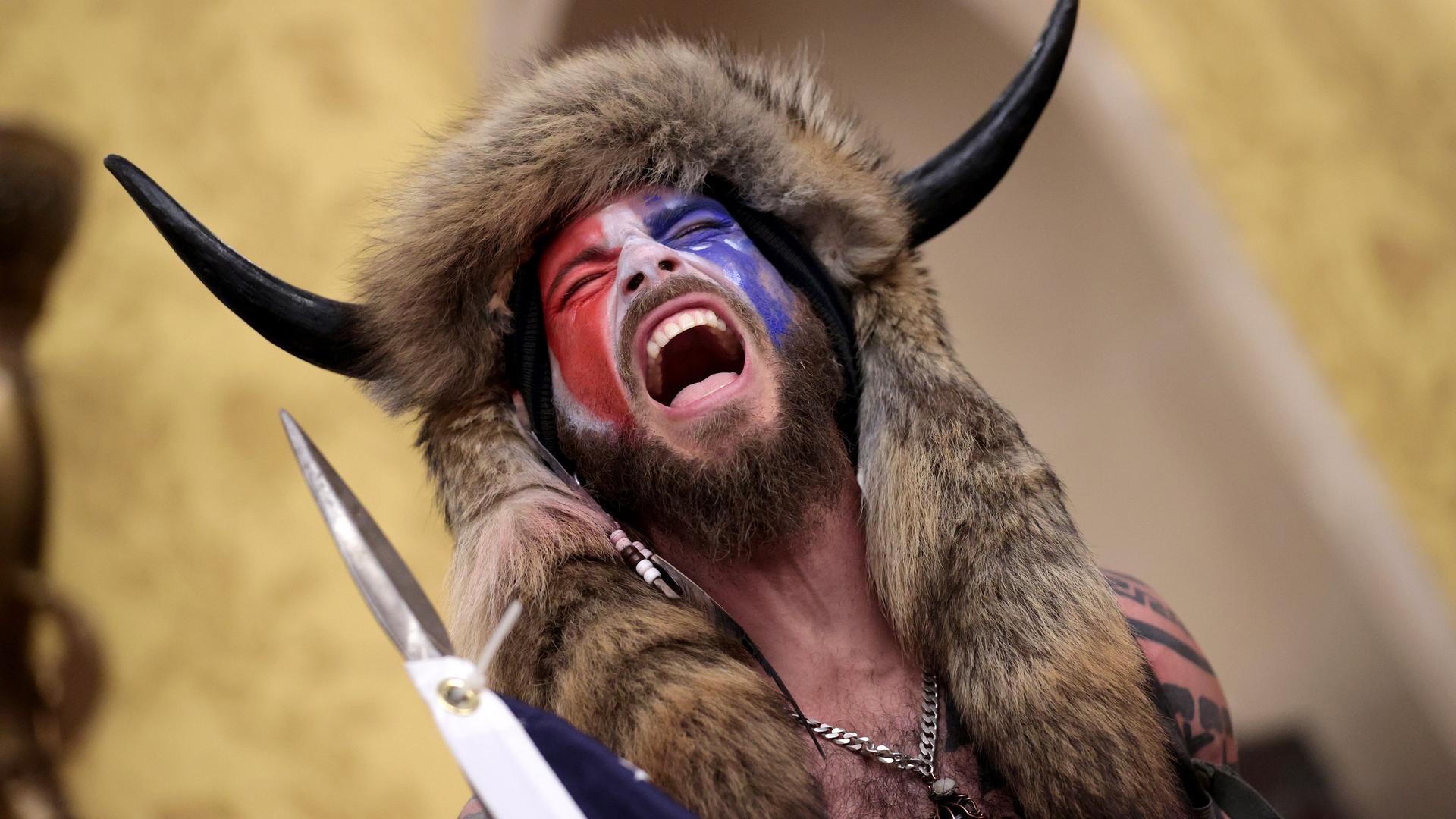 Ein Mann, dessen Gesicht mit der amerikanischen Flagge bemalt ist und der ein Tierfell und Hörner am Kopf trägt steht schreiend im Kaptitol