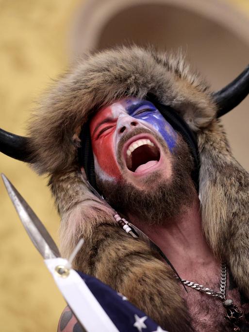 Ein Mann, dessen Gesicht mit der amerikanischen Flagge bemalt ist und der ein Tierfell und Hörner am Kopf trägt steht schreiend im Kaptitol
