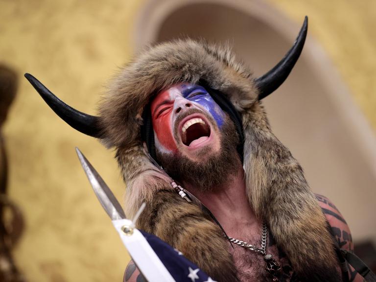 Ein Mann, dessen Gesicht mit der amerikanischen Flagge bemalt ist und der ein Tierfell und Hörner am Kopf trägt steht schreiend im Kaptitol