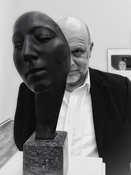 Der Fotokünstler Gregor Wildförster schaut hinter der Skulptur eines Gesichtes hervor. Sein Gesicht ist auf der Schwarz-Weiß-Fotografie dabei nur zur Hälfte zu sehen. 