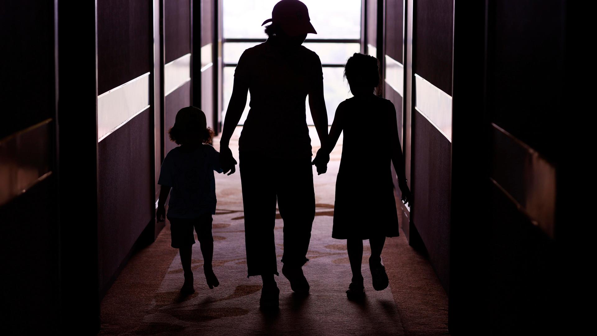 Silhouetten zweier Kinder, die an der Hand einer erwachsenen Person laufen. Im Gegenlicht sind lediglich ihre Silhouetten zu sehen. 