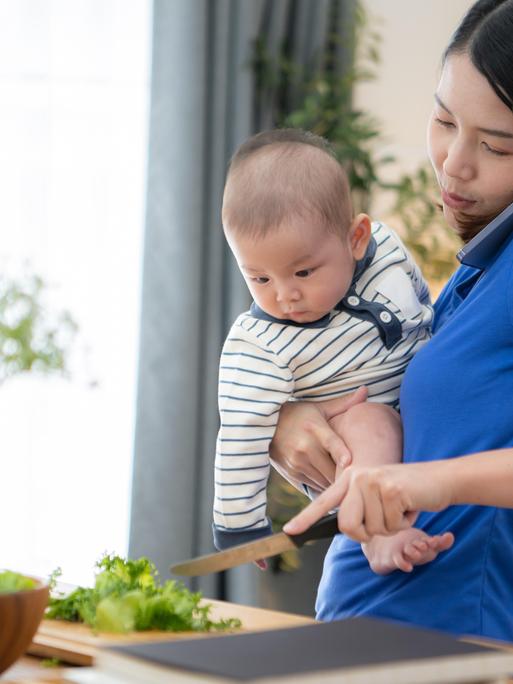 Mutter mit Handy am Ohr und Kleinkind auf dem Arm bereitet in der Küche das Abendessen zu