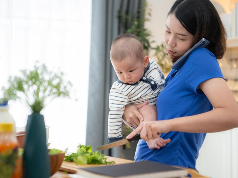 Mutter mit Handy am Ohr und Kleinkind auf dem Arm bereitet in der Küche das Abendessen zu