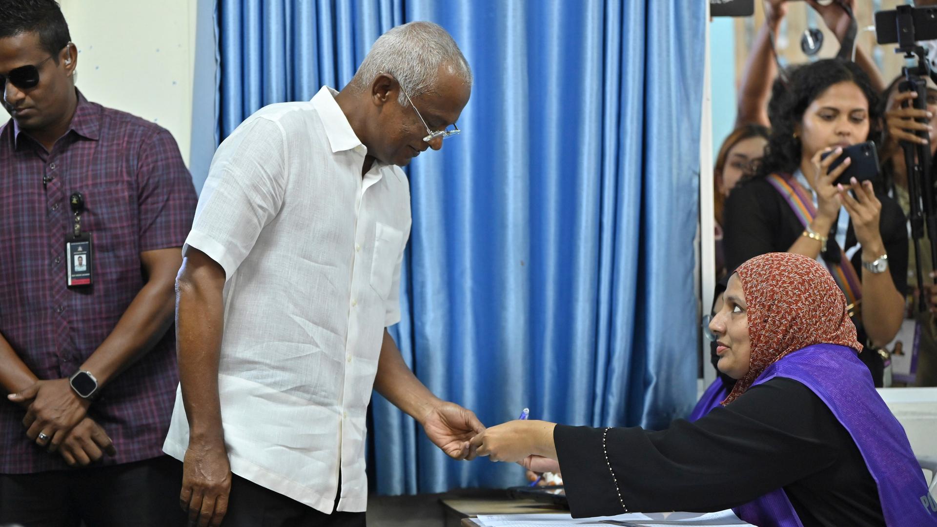 Solih im weißen Hemd steckt seinen Wahlzettel in eine Urne. An einem Tisch sitzt eine Wahlhelferin mit Kopftuch, die ihn lächelnd ansieht.