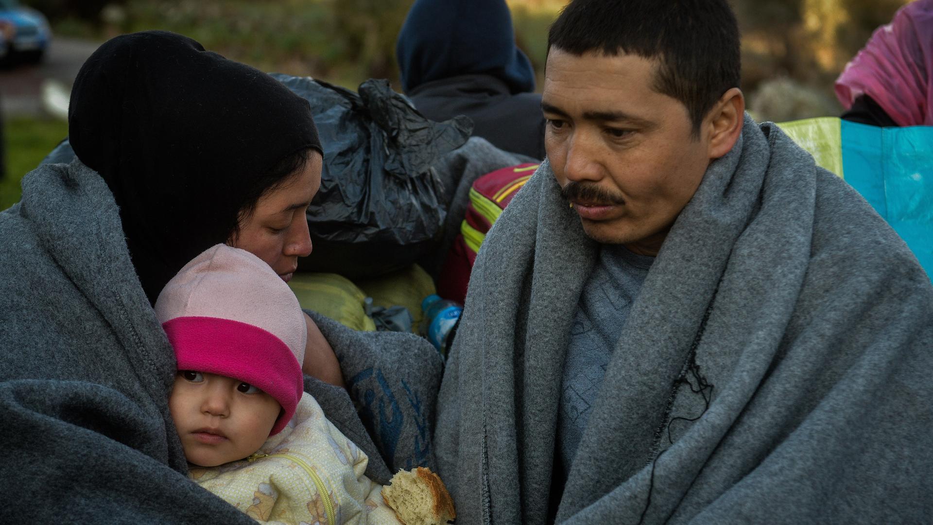 Eine geflüchtete Familie auf der Insel Lesbos in Griechenland. Mutter und Vater haben graue Decken umgelegt. Die Mutter hält ein kleines Kind im Arm.