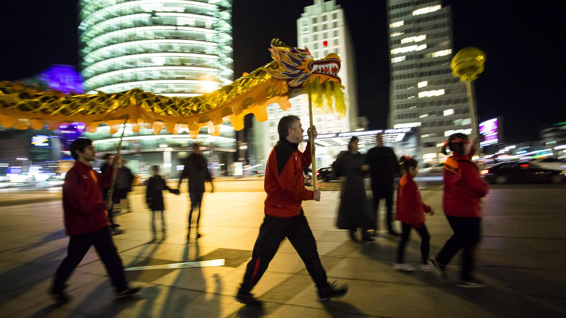 Drachentanz zum chinesischen Neujahr am Potsdamer Platz in Berlin im Jahr 2018: Mehrere Menschen in roten Jacken tragen an Stäben eine Drachenfigur und eine goldene Kugel vor der Kulisse erleuchteter Hochhäuser.  