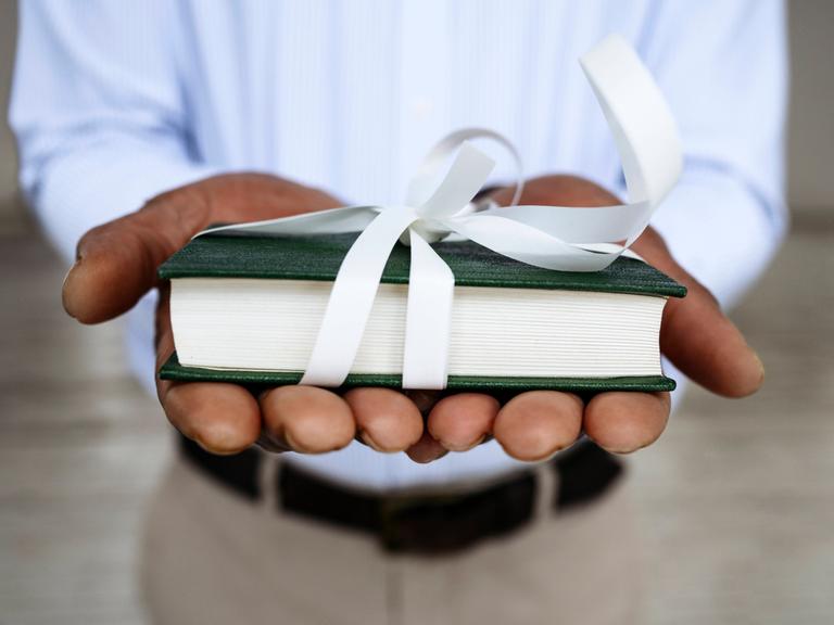 Nahaufnahme zweier Hände, die der Kamera ein mit weißem Geschenkband verziertes Buch entgegenhalten.