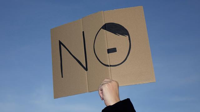 Jemand hält ein Pappschild in die Luft, auf dem "NO" geschrieben steht. Der Buchstabe "O" wurde mit einen Seitenscheitel und einem Bärtchen versehen, sodass er an Hitler erinnert. 