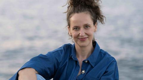 Carolin Würfel sitzt mit hochgesteckten Haaren an einem See und schaut freundlich in die Kamera.