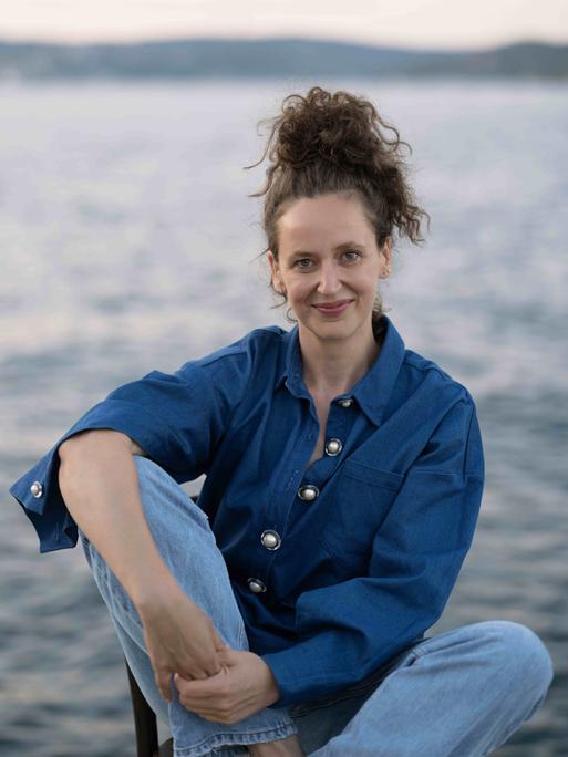 Carolin Würfel sitzt mit hochgesteckten Haaren an einem See und schaut freundlich in die Kamera.