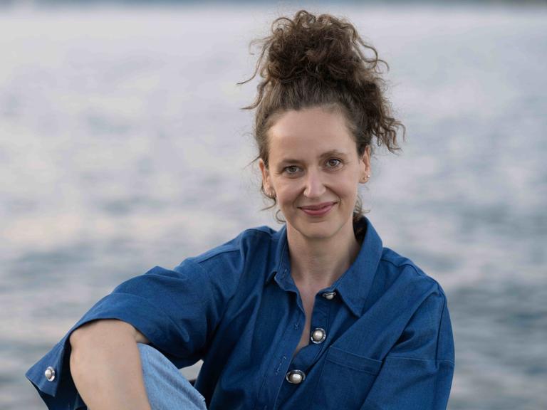 Carolin Würfel sitzt mit hochgesteckten Haaren an einem See und schaut freundlich in die Kamera.