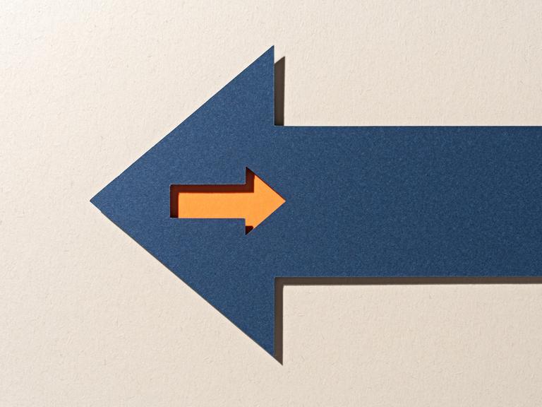 Ein orangefarbener kleiner Pfeil in einem großen blauen Pfeil, der sich in umgekehrter Richtung auf beigem Hintergrund bewegt.