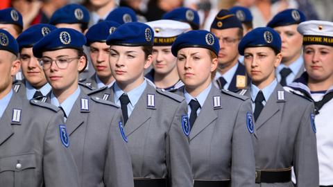 Soldatinnenund Soldaten stehen in mehreren Reihen hinterinander in grauen Uniformen und blauen Baretten. Dazwischen ein Soldat und eine Soldatin der Marine in weiß.