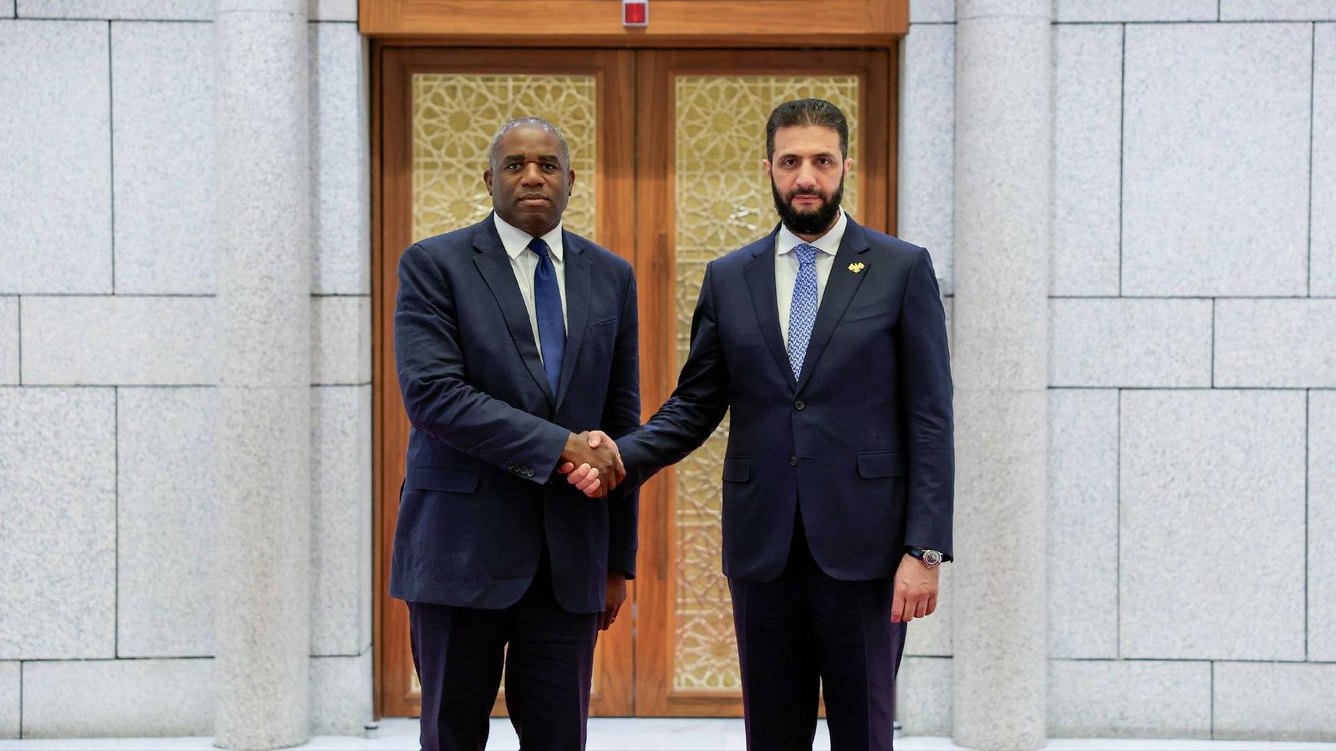 Ahmed al-Sharaa und David Lammy schütteln sich die Hand und schauen in die Kamera.