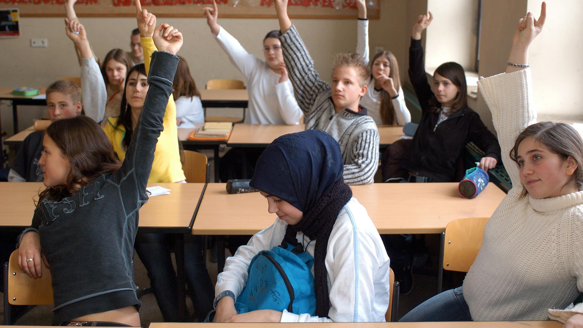 Mehrere Kinder sitzen in einer Bank-Reihe. Einige heben die Hand und melden sich. Ein Mädchen trägt ein Kopf-Tuch.