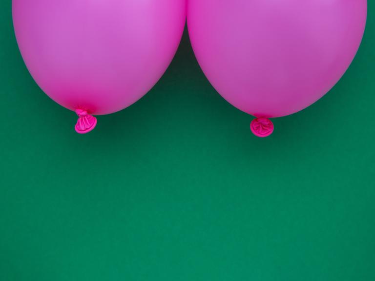 Zwei pinke Luftballons im Anschnitt, die zwei Brüste symbolisieren vor grünem Hintergrund.