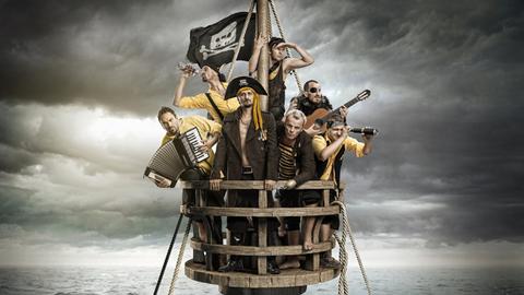 Blick auf die Band, die sich in Piratenkostümen auf einem Mastkorb befinden und Ausschau halten, während ihr Schiff schon untergegangen ist und Haie um den Mast kreisen. Dabei haben sie diverse Instrumente in den Händen. Blick auf die Band, die sich in Piratenkostümen auf einem Mastkorb befinden und Ausschau halten, während ihr Schiff schon untergegangen ist und Haie um den Mast kreisen. Dabei haben sie diverse Instrumente in den Händen.