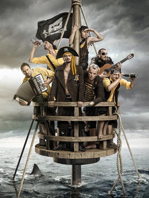 Blick auf die Band, die sich in Piratenkostümen auf einem Mastkorb befinden und Ausschau halten, während ihr Schiff schon untergegangen ist und Haie um den Mast kreisen. Dabei haben sie diverse Instrumente in den Händen.