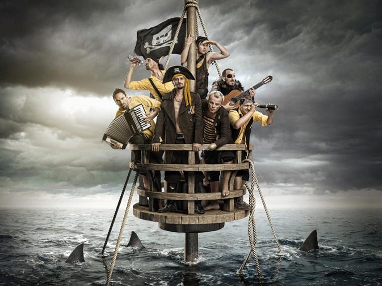 Blick auf die Band, die sich in Piratenkostümen auf einem Mastkorb befinden und Ausschau halten, während ihr Schiff schon untergegangen ist und Haie um den Mast kreisen. Dabei haben sie diverse Instrumente in den Händen.