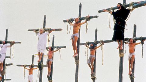 Szene aus Monty Phytons "Leben des Brian" bei der Kreuzigung Jesu Christi. Mehrere Männer in weißen oder schwarzen Gewändern hängen an Kreuzen. 