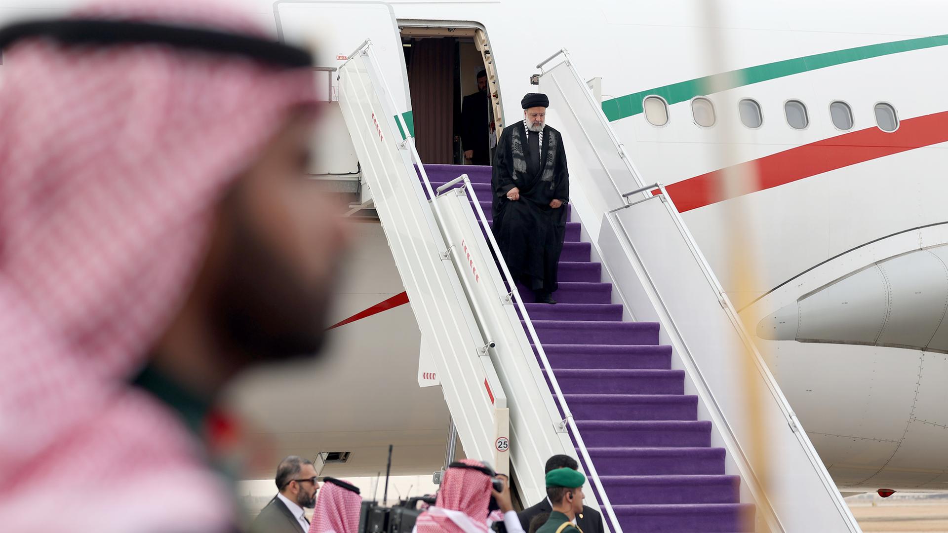 Raisi in schwarzer Kleidung schreitet die mit lila Stoff ausgelegte Treppe von seinem Flugzeug herunter, anderen Ende das Begrüßungskomitee wartet. Im Vordergrund unscharf der Kopf eines bärtigen Mannes mit weiß-roter Kopfbedeckung. Raisi in schwarzer Kleidung schreitet die mit lila Stoff ausgelegte Treppe von seinem Flugzeug herunter, anderen Ende das Begrüßungskomitee wartet. Im Vordergrund unscharf der Kopf eines bärtigen Mannes mit weiß-roter Kopfbedeckung.