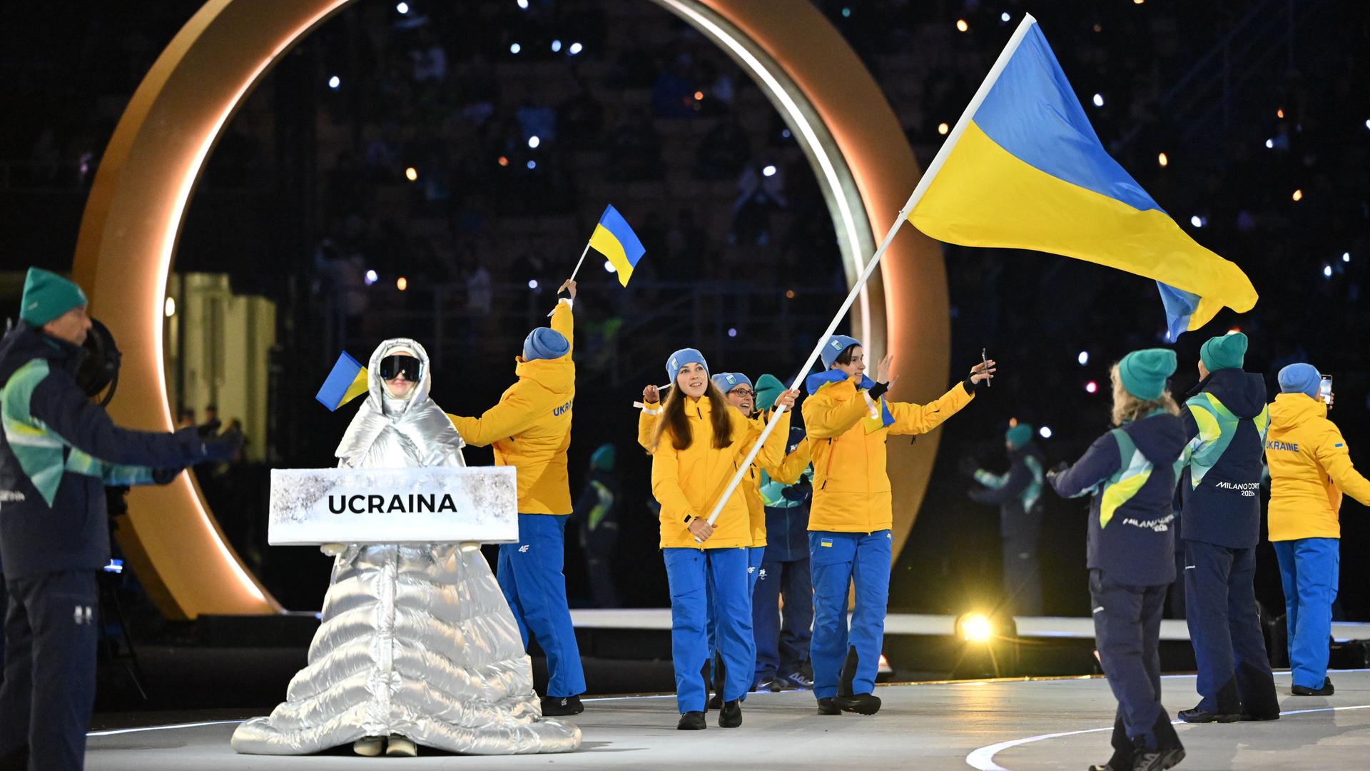 Sportlerinnen und Sportler aus der Ukraine stehen auf der Bühne. Sie tragen Schnee-Anzüge in gelb und blau. Vor ihnen steht eine Frau in einem silbernen Kostüm. Sie trägt ein Schild mit der Aufschrift "Ukraine".