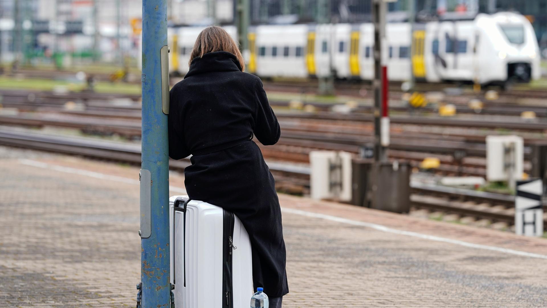 Eine Frau sitzt an einem Bahnsteig auf ihrem Koffer und wartet.