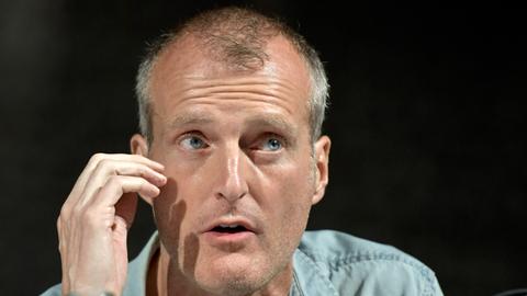 Nahaufnahme eines Gesichtes eines Mannes mit kurzem grauem Haar und weit geöffneten blauen Augen. Er trägt ein blaues Jeanshemd und spricht vor schwarzem Hintergrund. Dazu gestikuliert er mit seiner rechten Hand vor dem Gesicht. 