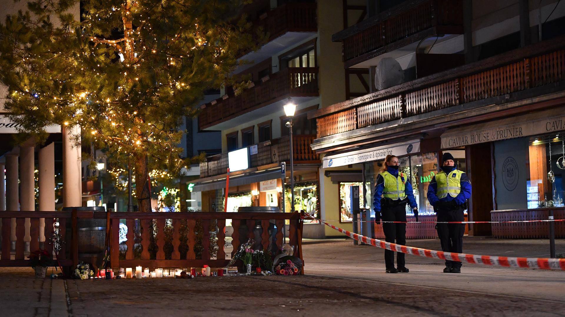 Kerzen und Blumen stehen vor einem Zaun im Ort Crans-Montana. Zwei Sicherheitskräfte stehen hinter einem Absperrband.