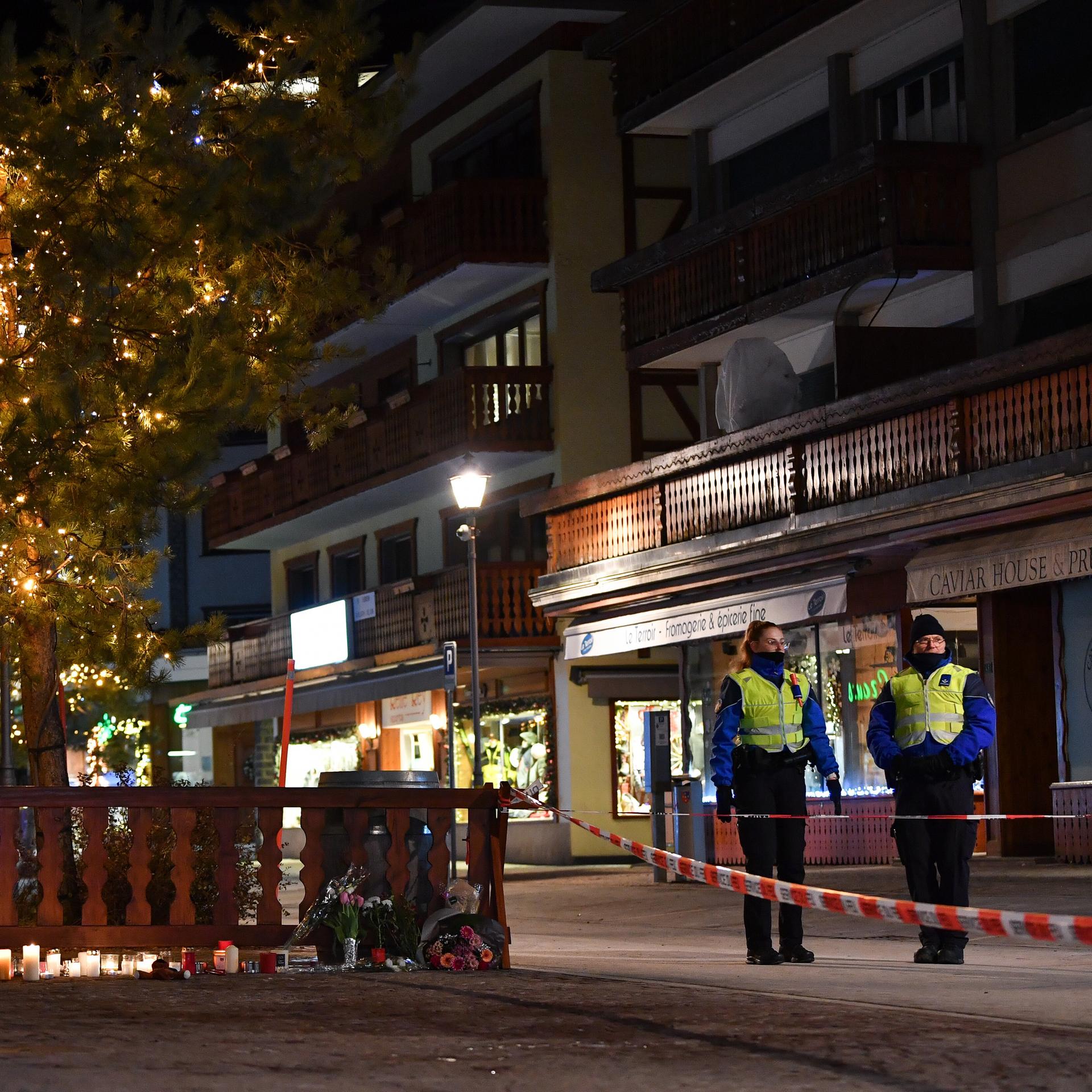 Kerzen und Blumen stehen vor einem Zaun im Ort Crans-Montana. Zwei Sicherheitskräfte stehen hinter einem Absperrband.