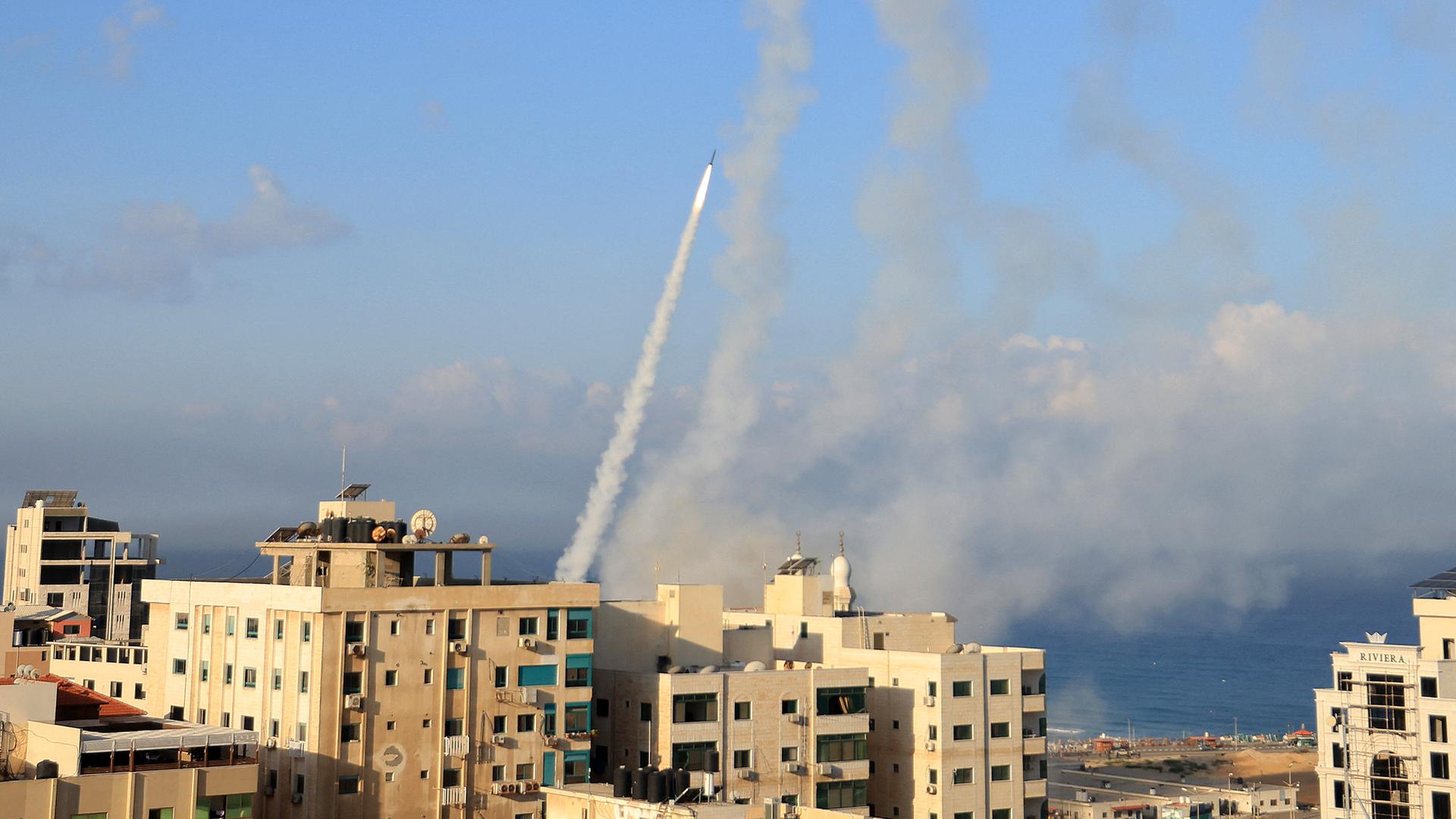 Das Bild zeigt Gaza-Stadt. Im Hintergrund erkennt man eine Rakete kurz nach dem Start.  Sandfarbene Mehrfamilienhäuser sind im Vordergrund zu sehen.