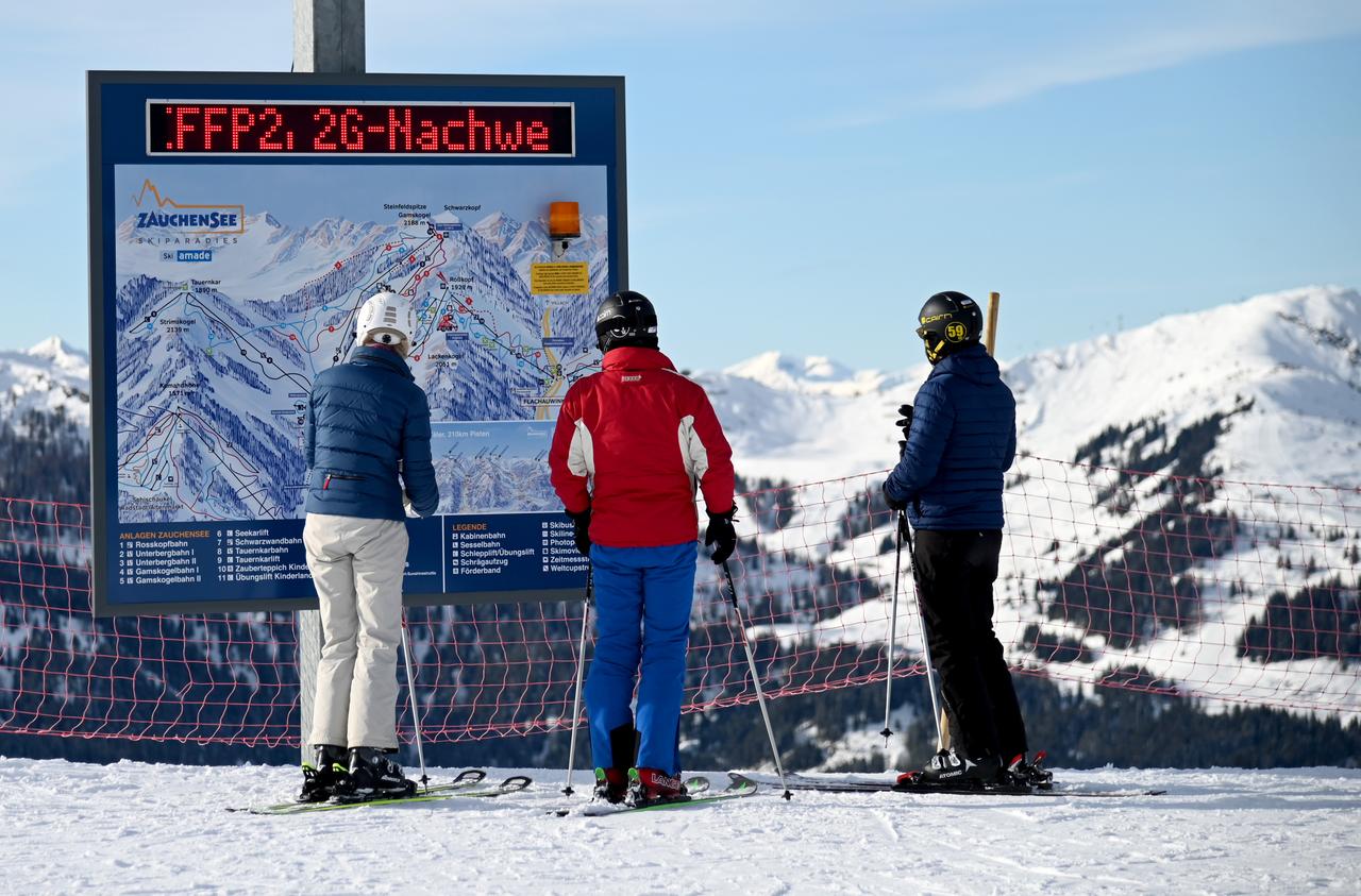 Skifahrer im österreichischen Zauchensee stehen vor einer Informationstafel mit der Aufforderung nach FFP2-Masken und 2G-Nachweis