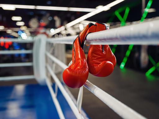 Zwei rote Boxhandschuhe hängen in den Seilen eines Boxrings.