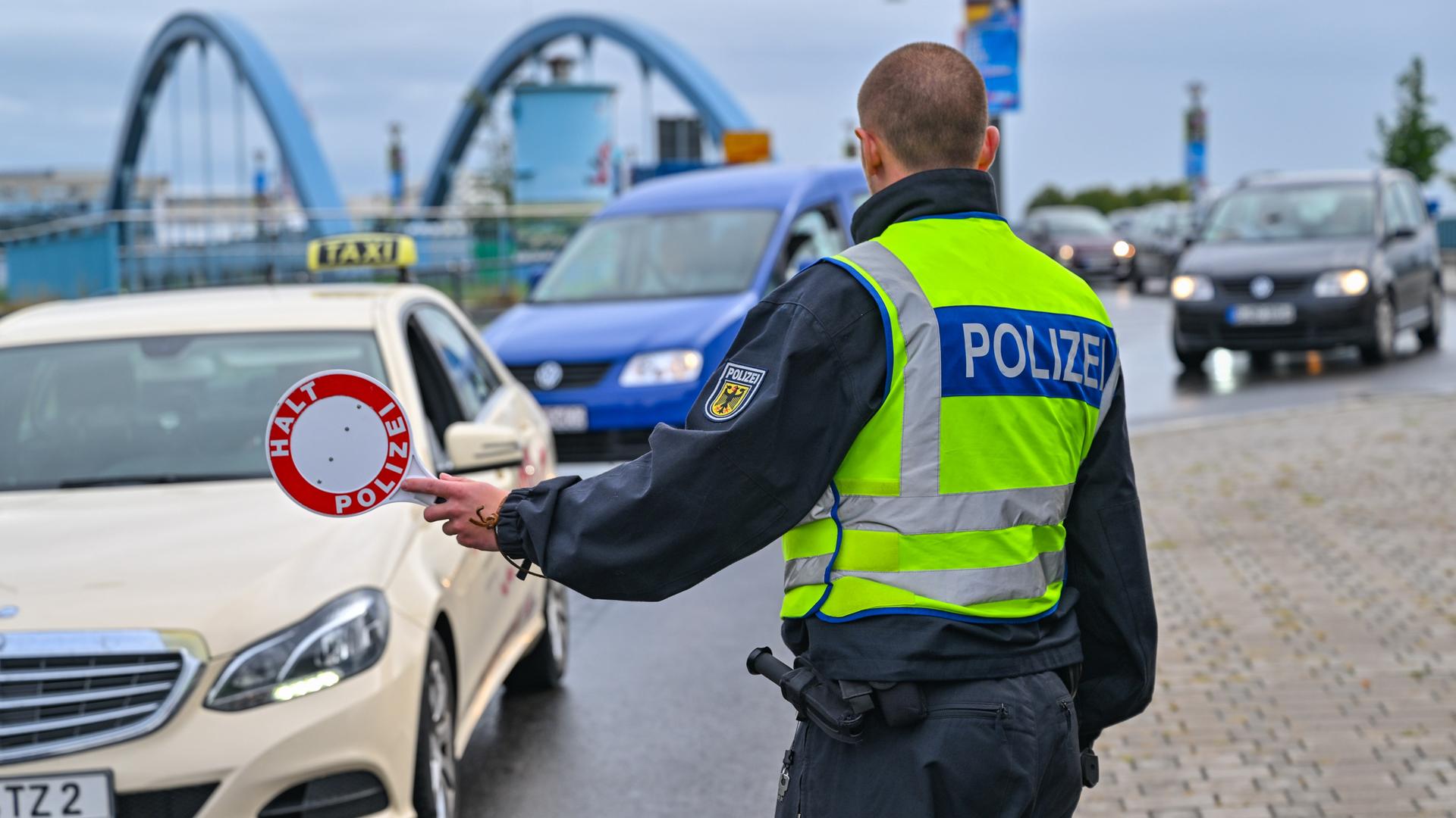 Ein Polizist hält Autos zur Kontrolle am Grenzübergang an. Im Hintergrund ist eine Brücke zu sehen.