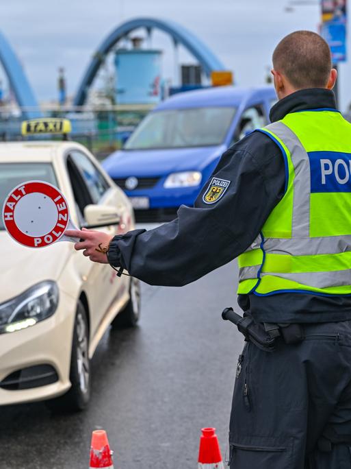 Ein Polizist hält Autos zur Kontrolle am Grenzübergang an. Im Hintergrund ist eine Brücke zu sehen.