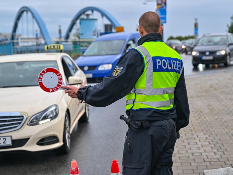 Ein Polizist hält Autos zur Kontrolle am Grenzübergang an. Im Hintergrund ist eine Brücke zu sehen.