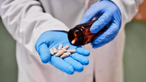 Nahaufnahme: Hand in blauem Hygienehandschuh hält Tabletten, die aus einem kleinen Glasbehälter geschüttet wurden.