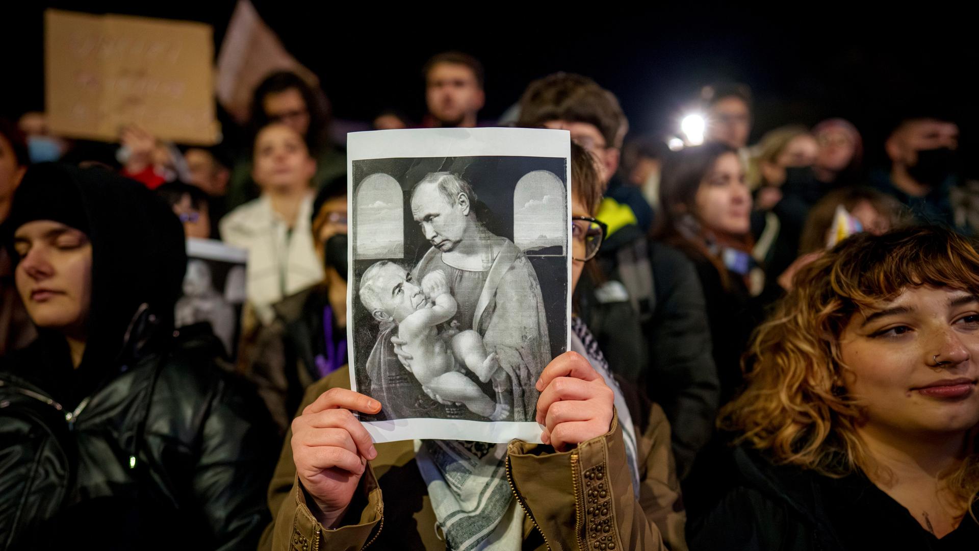 Demonstranten stehen auf einem Platz in Rumänien, eine Person in der Mitte hält ein satirisches Poster. Es zeigt Calin Georgescu, der an der nährenden Brust von Russlands Präsident Wladimir Putin saugt.