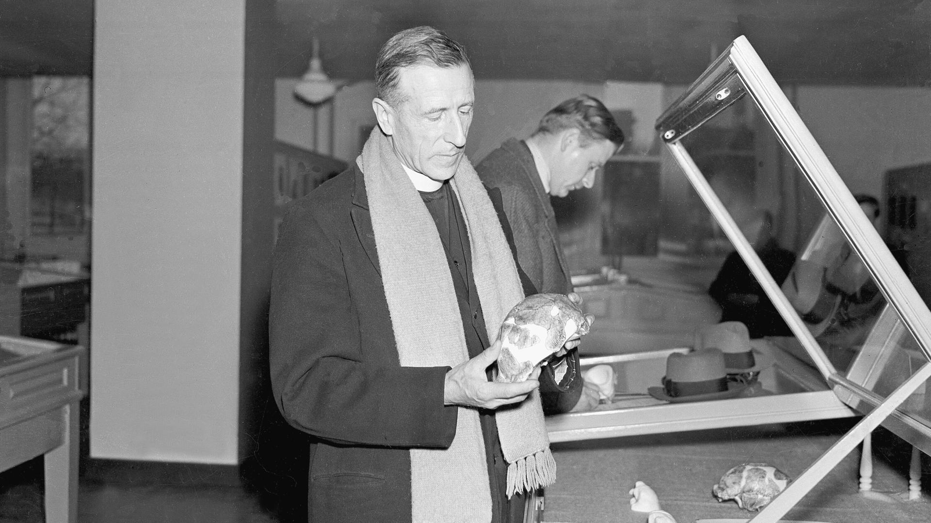 Historisches Schwarzweißfoto von Pierre Teilhard de Chardin, der vor einem Schaukasten steht und einen menschlichen Schädel in der Hand hält. Historisches Schwarzweißfoto von Pierre Teilhard de Chardin, der vor einem Schaukasten steht und einen menschlichen Schädel in der Hand hält.