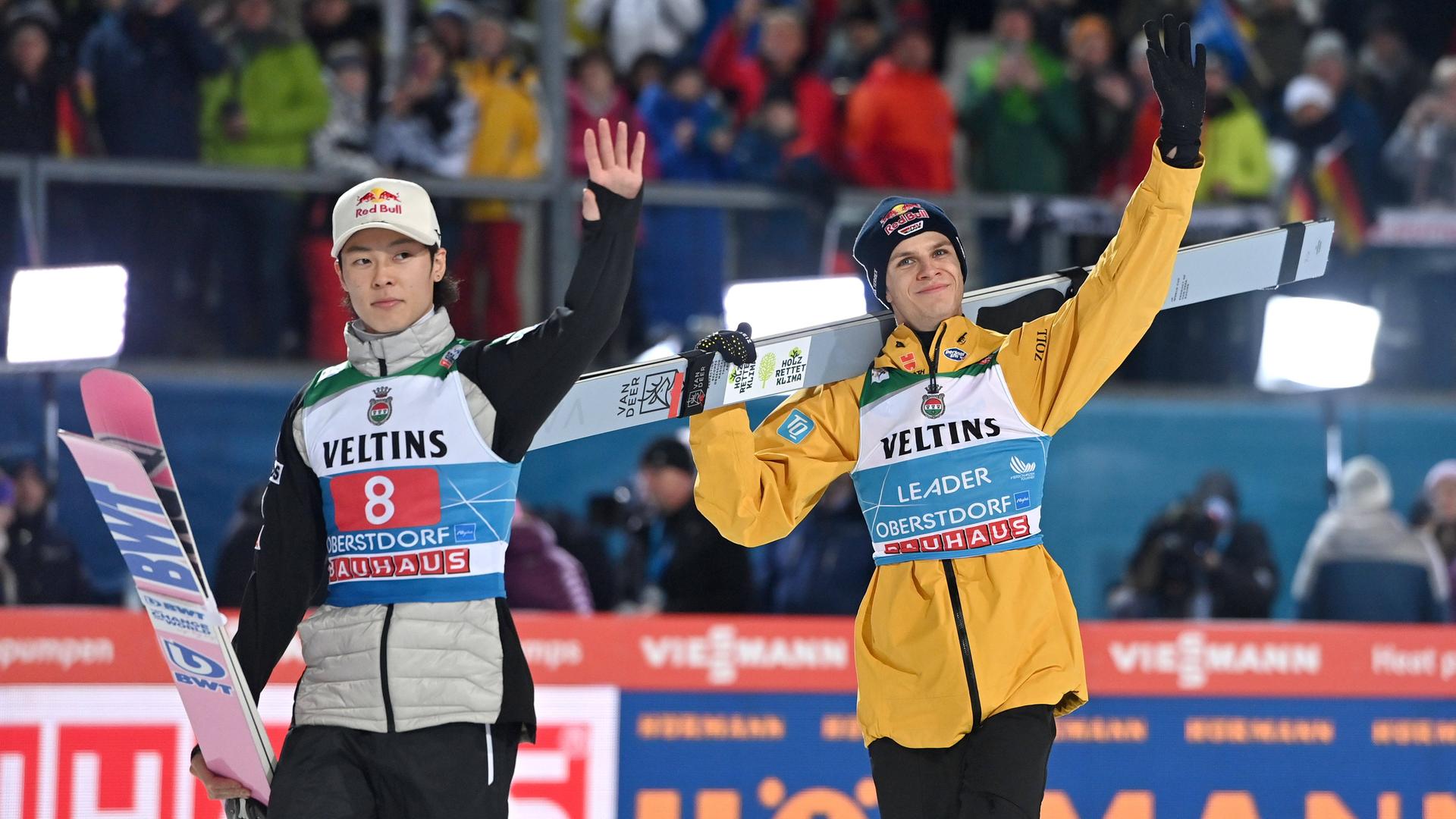 Die Skispringer Andreas Wellinger (r.) und Ryoyu Kobayashi tragen ihre Skier und winken ins Publikum.