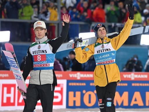Die Skispringer Andreas Wellinger (r.) und Ryoyu Kobayashi tragen ihre Skier und winken ins Publikum.