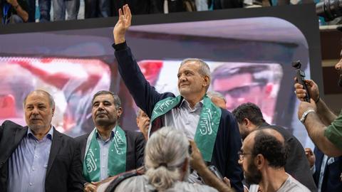 Massud Peseschkian ist ein Mann mit kurzen grauen Haaren. Er steht zwischen anderen Männern und hebt die Hand.