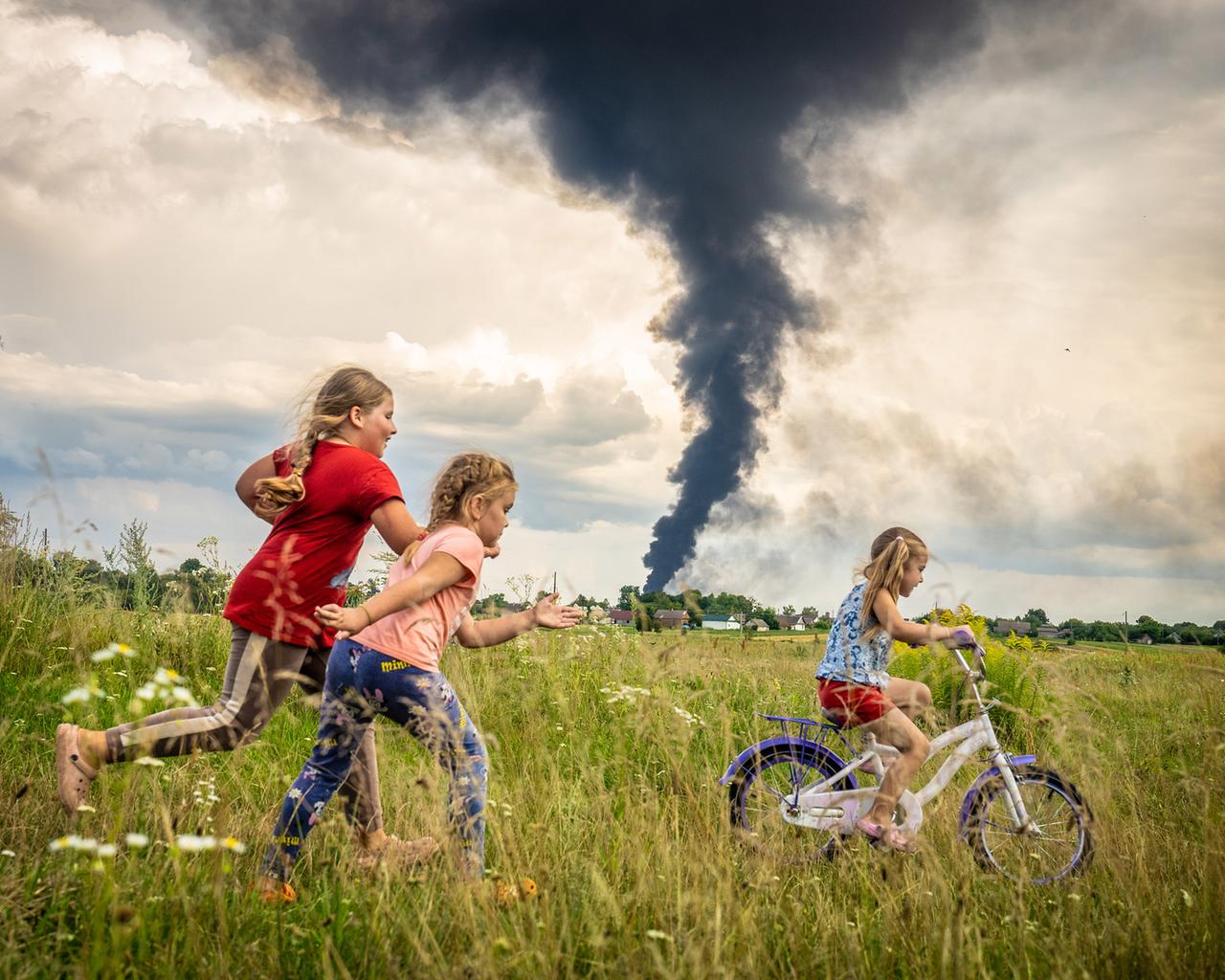 Das Foto zeigt drei Kinder auf einer Wiese, eines davon auf einem Fahrrad. Im Hintergrund steigt eine Rauchsäule auf.