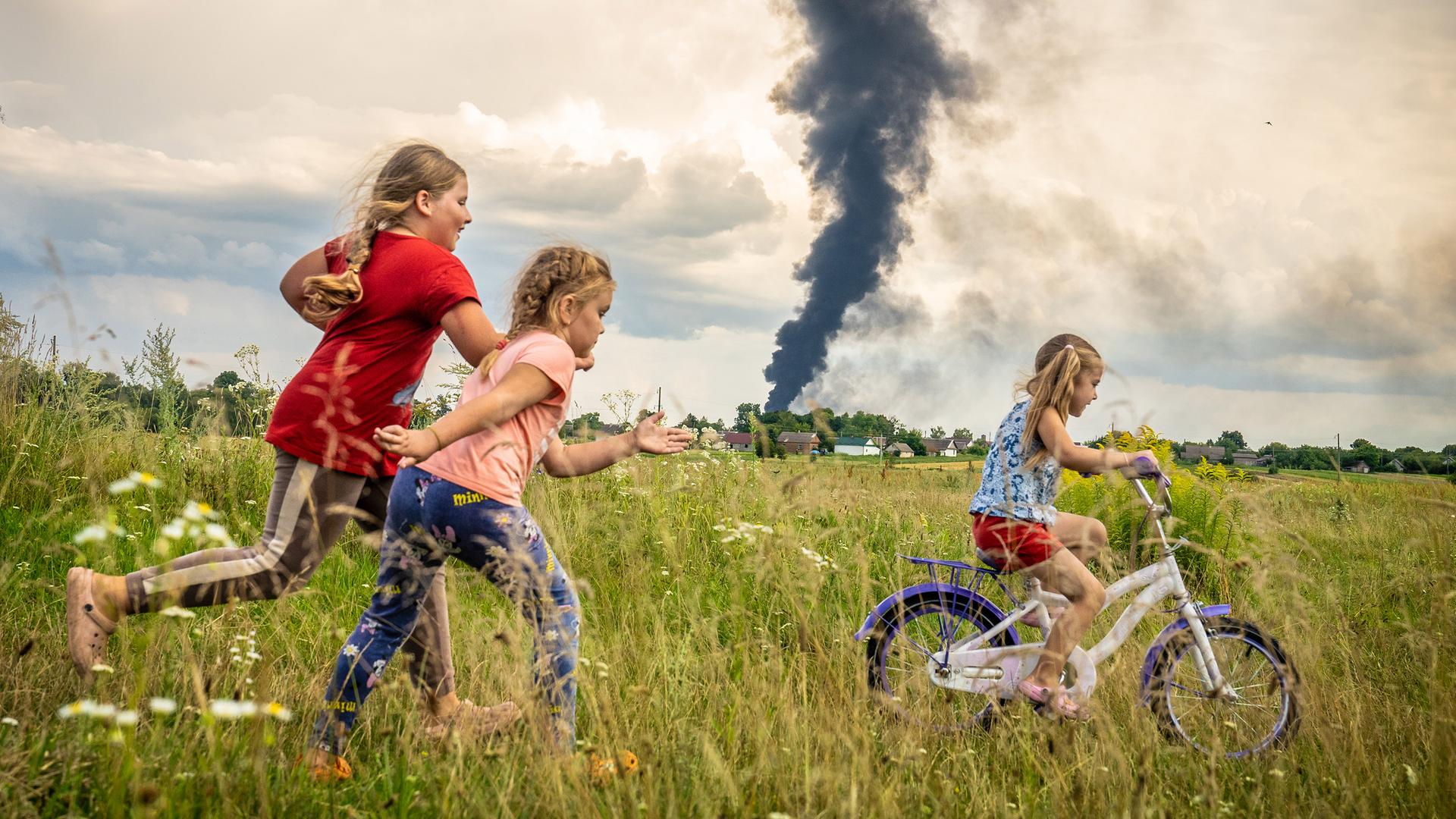 Das Foto zeigt drei Kinder auf einer Wiese. Ein Kind fährt Fahrrad. Im Hinter-Grund steigt eine Rauch-Säule auf.