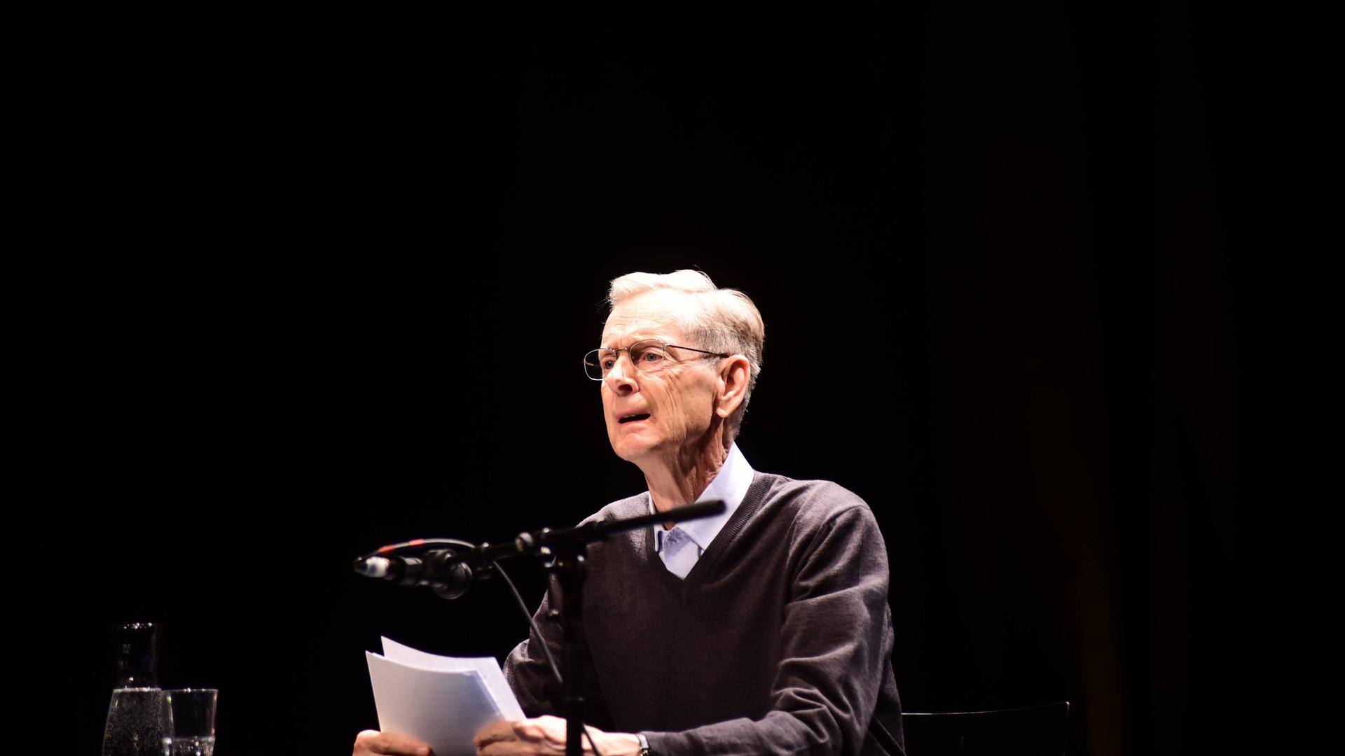 Walter Kappacher 2017 bei einer Lesung. Er sitzt an einem Tisch vor einem Mikrophon und hält Papierblätter in der Hand.
