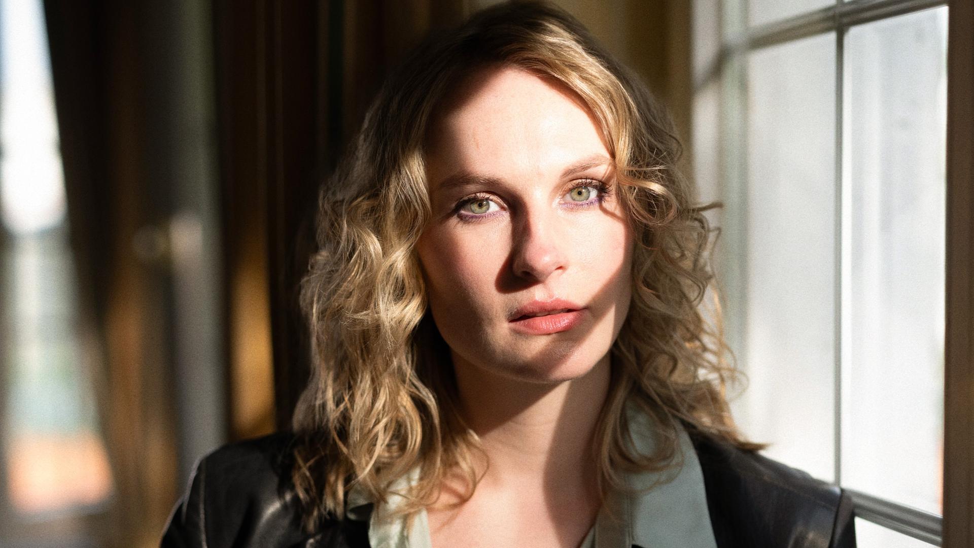 Porträt der Drehbuchautorin und Regisseurin Marie C. König. Sie steht neben einem Fenster, das Licht fällt auf ihre blonden Haare. Porträt der Drehbuchautorin und Regisseurin Marie C. König. Sie steht neben einem Fenster, das Licht fällt auf ihre blonden Haare.