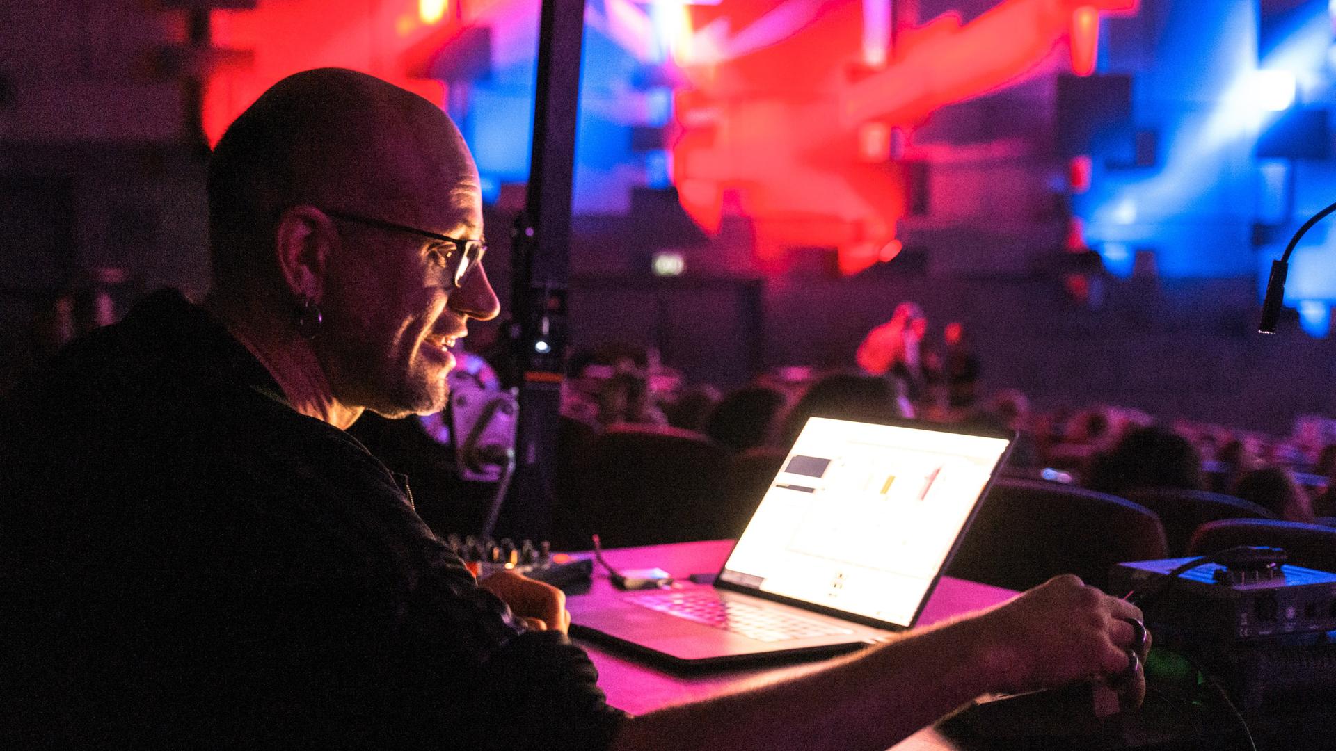Der Medienkünstler Robert Henke schaut konzentriert auf einen Laptop und dreht an Reglern, der ganze Raum ist in rotes und blaues Licht getaucht.