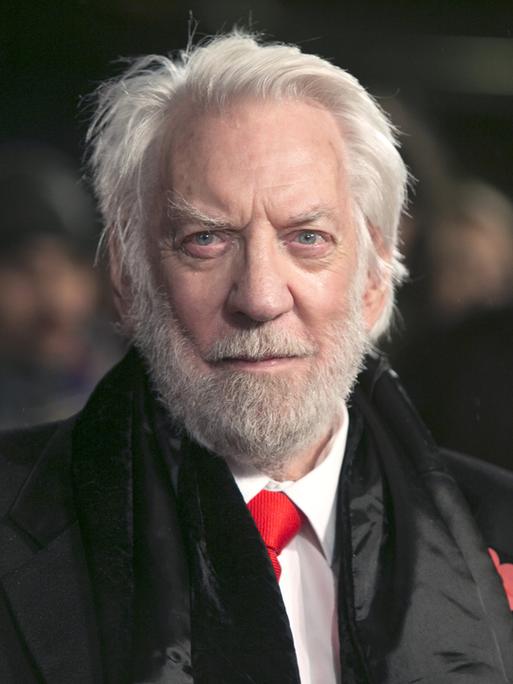 Donald Sutherland trägt einen schwarzen Mantel, ein weißes Hemd, eine rote Krawatte und schaut in die Kamera.