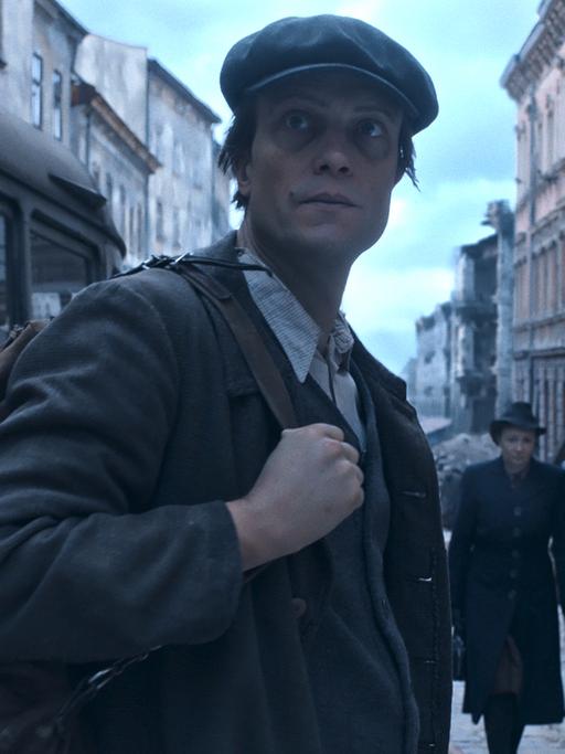 Eine Straßenszene in einer zerstörten deutschen Stadt nach dem 2. Weltkrieg. Ein Mann mit Rucksack blickt in die Kamera, zwei Frauen gehen an ihm vorbbei, Männer mit Schaufeln räumen Schutt weg.